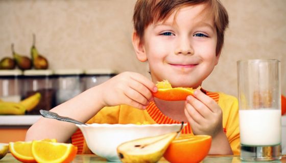 Grinsender Junge isst ein Stück Orange, vor sich eine Müslischale, drumherum Obst