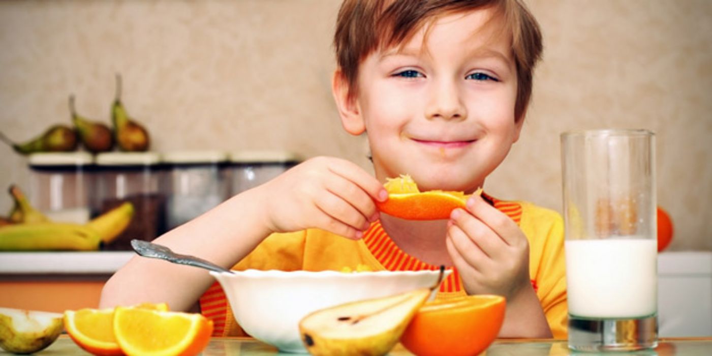 Grinsender Junge isst ein Stück Orange, vor sich eine Müslischale, drumherum Obst