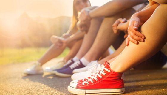 Nackte Beine von jungen Frauen die am Boden sitzen in Abendlicht, alle mit Sneakers (Chucks?), im Vordergrund rote mit weißen Sohlen