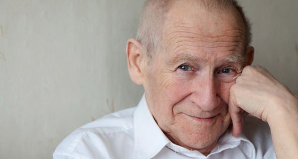 Portrait Senior, ca. Anfang 70, linke Hand stützt Wange, schütteres, etwas rötliches Haar, schaut verschmitzt lächelnd in die Kamera. Weißer Hemdkragen