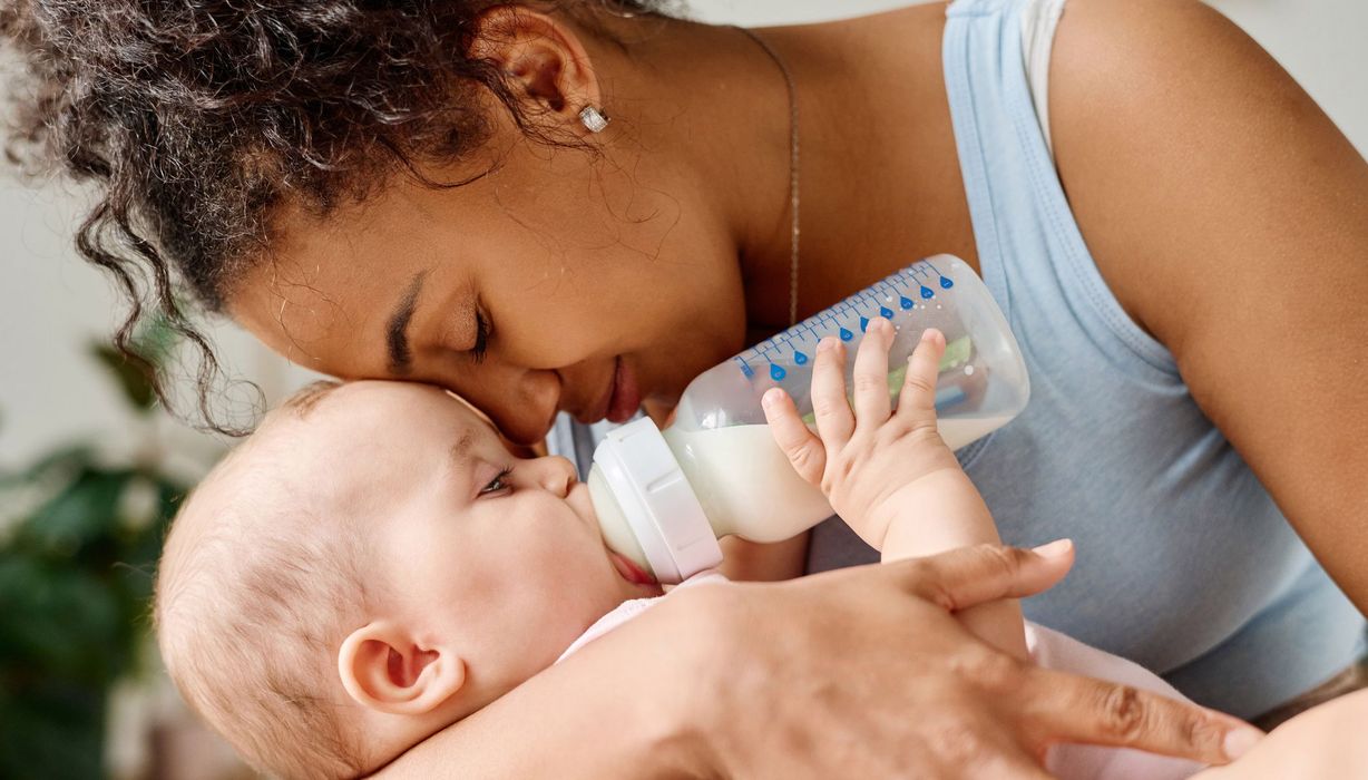 Baby trinkt aus einer Flasche, während eine Frau mit ihm kuschelt.