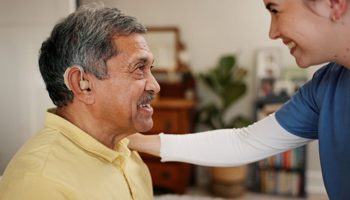 Ein älterer Mann mit einem Hörgerät und eine junge Frau lächeln sich an. 