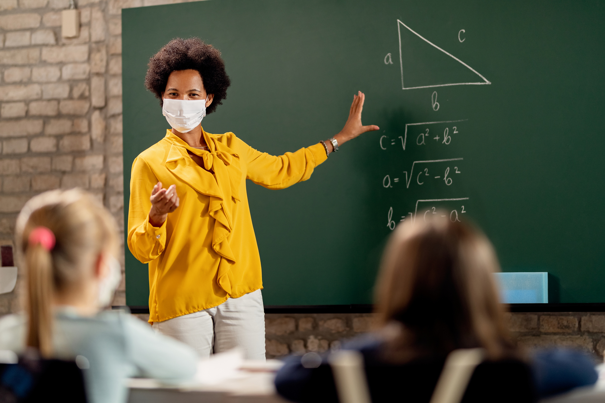 Lehrerin mit Maske steht an der Tafel und erklärt ihren Schülern eine Matheaufgabe.